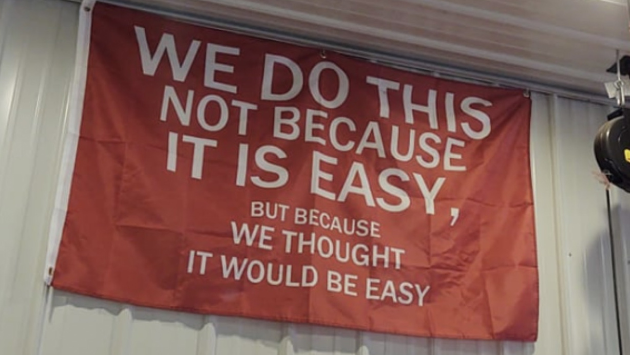 A red banner on a corrugated metal wall reading, in bold white capitals, "We do this not because it is easy, but because we thought it would be easy" — a play on the JFK moon-speech line.